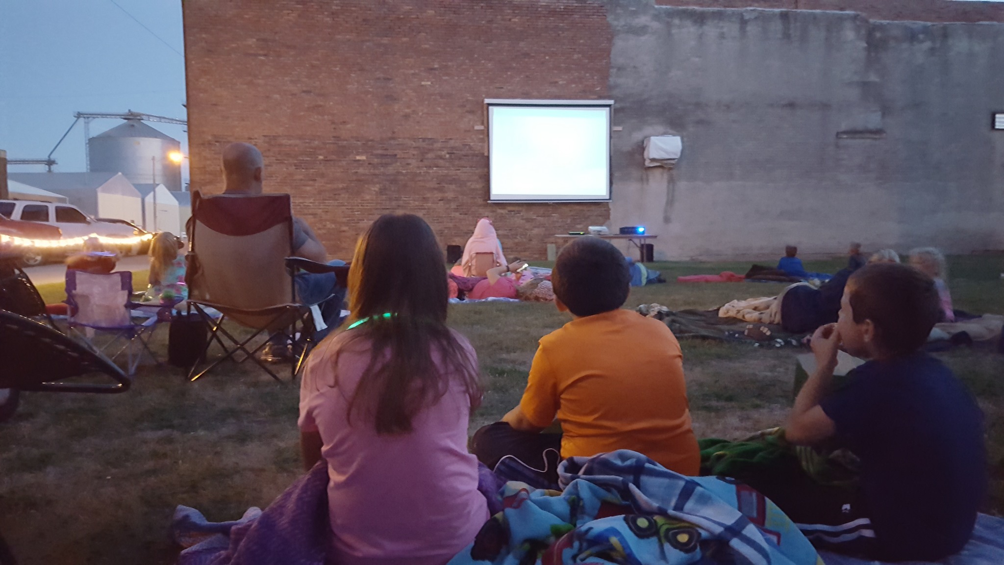 A group of people watching a movie outdoors on a video screen on the side of a brick building.