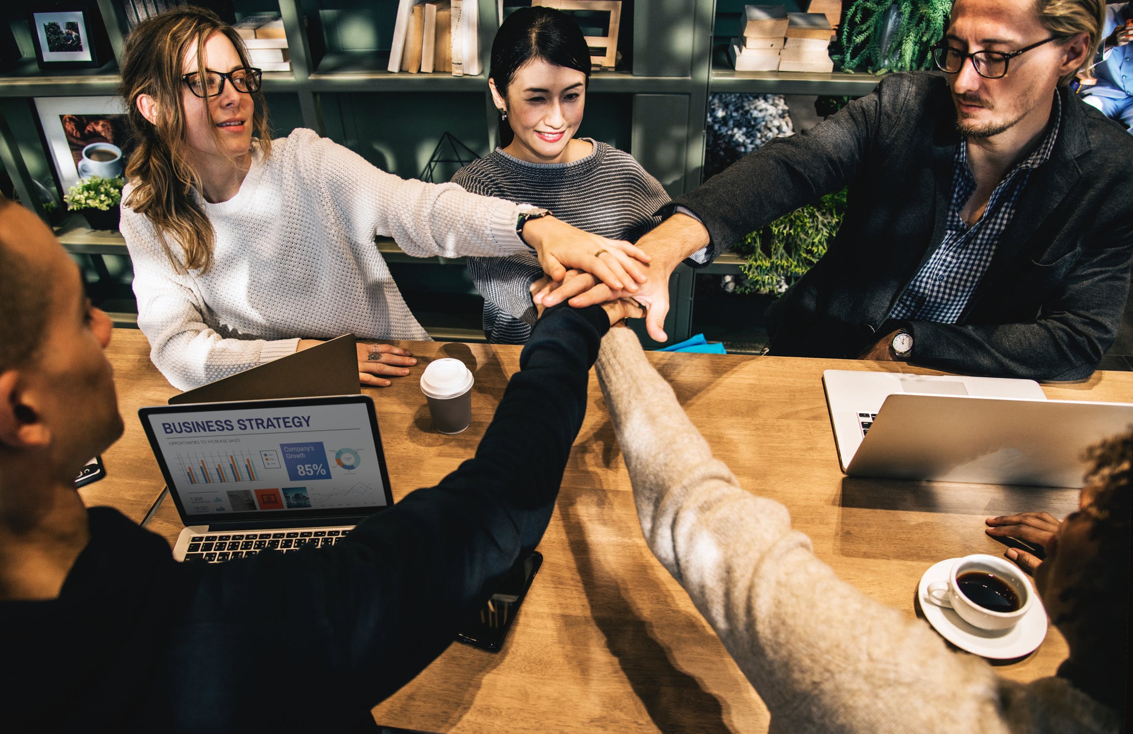 A group of young workers sitting at a table putting their hands together.