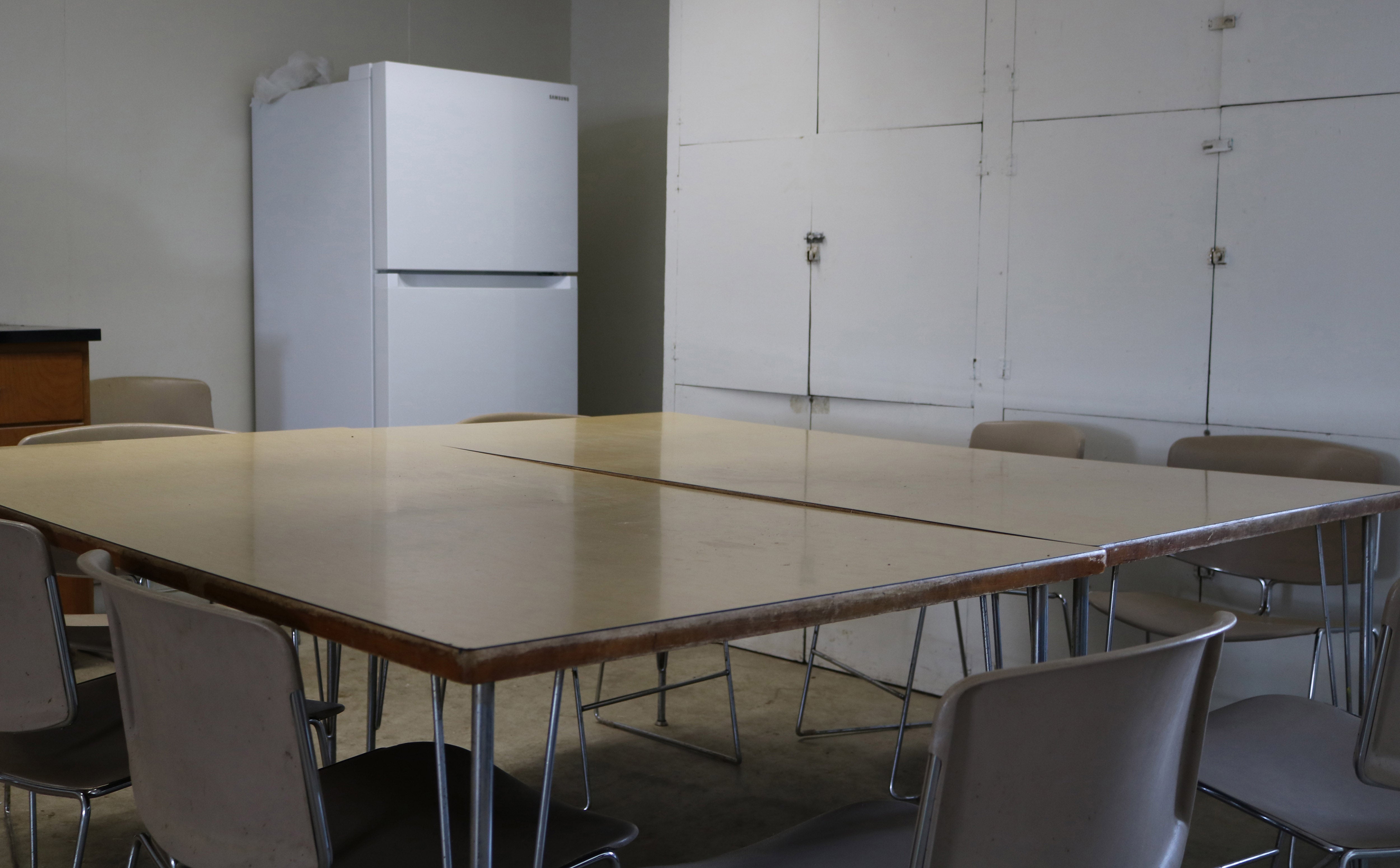 large table with several chairs around it. a refrigerator is in the background.