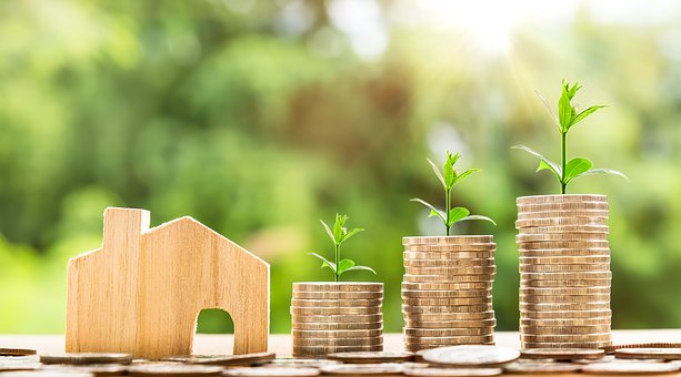 A small, wooden house cutout with three stacks of coins to the right. The stacks are get larger as you go right.