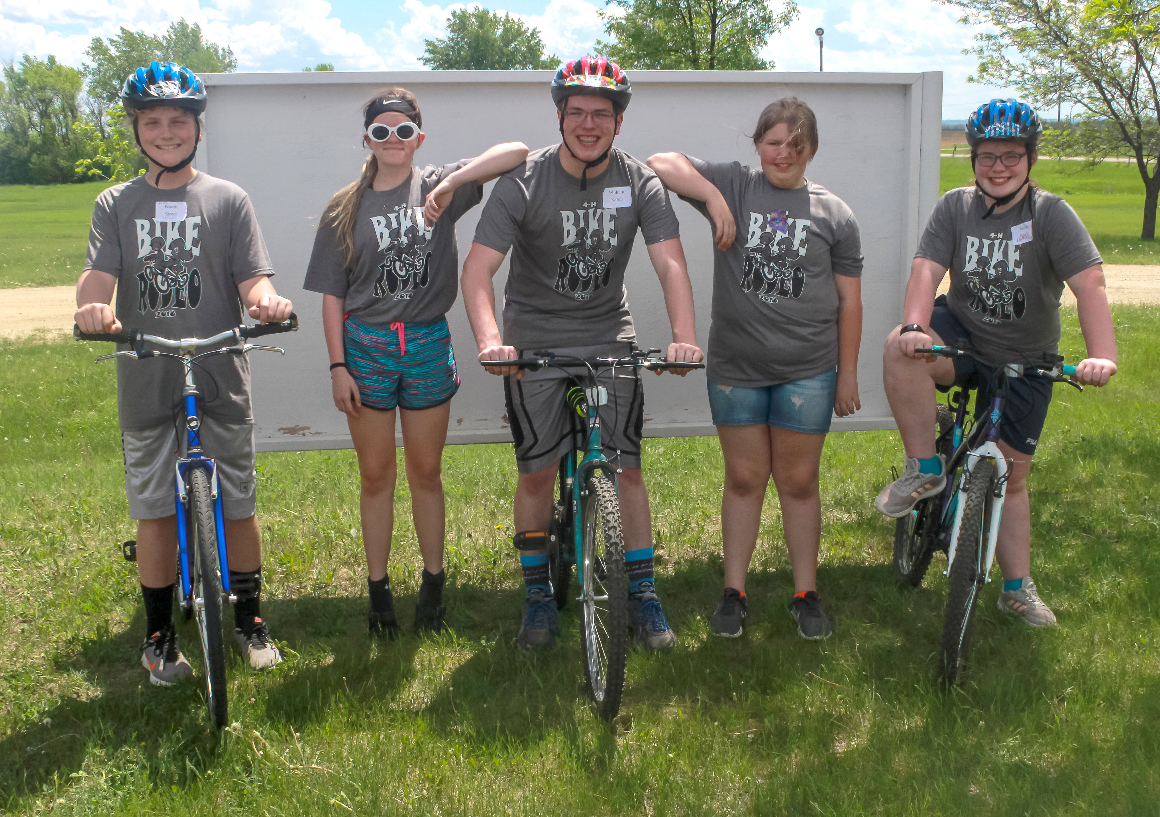 a group of five 4-H youth. two males and one female are on bicycles. two female youth are standing in the background.