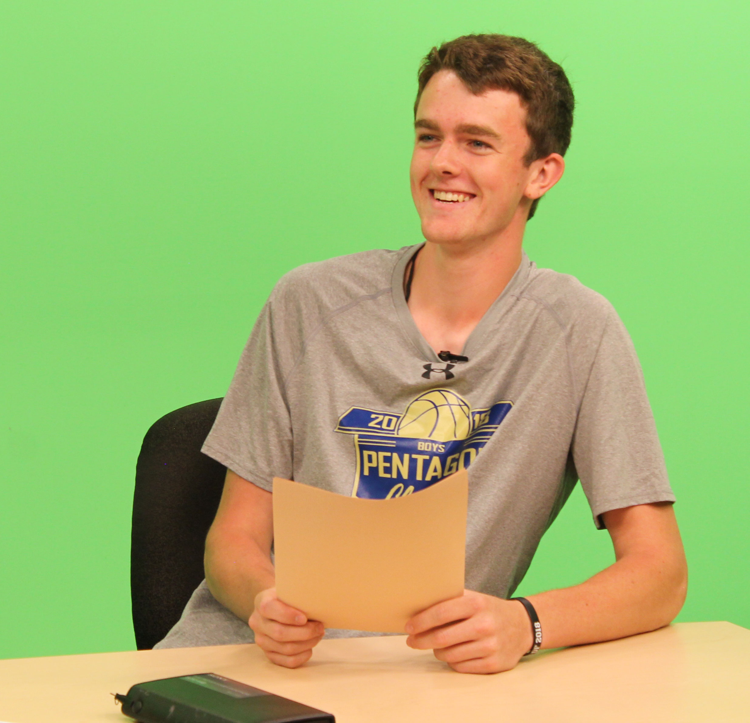 a teenage male sitting at a news desk in front of a green screen