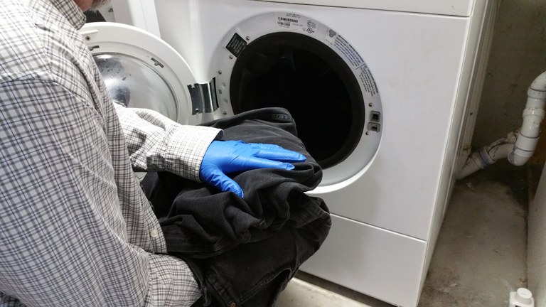 Man wearing a long-sleeved shirt and waterproof gloves loading pesticide-contaminated clothing into a washer. Courtesy: NDSU Extension
