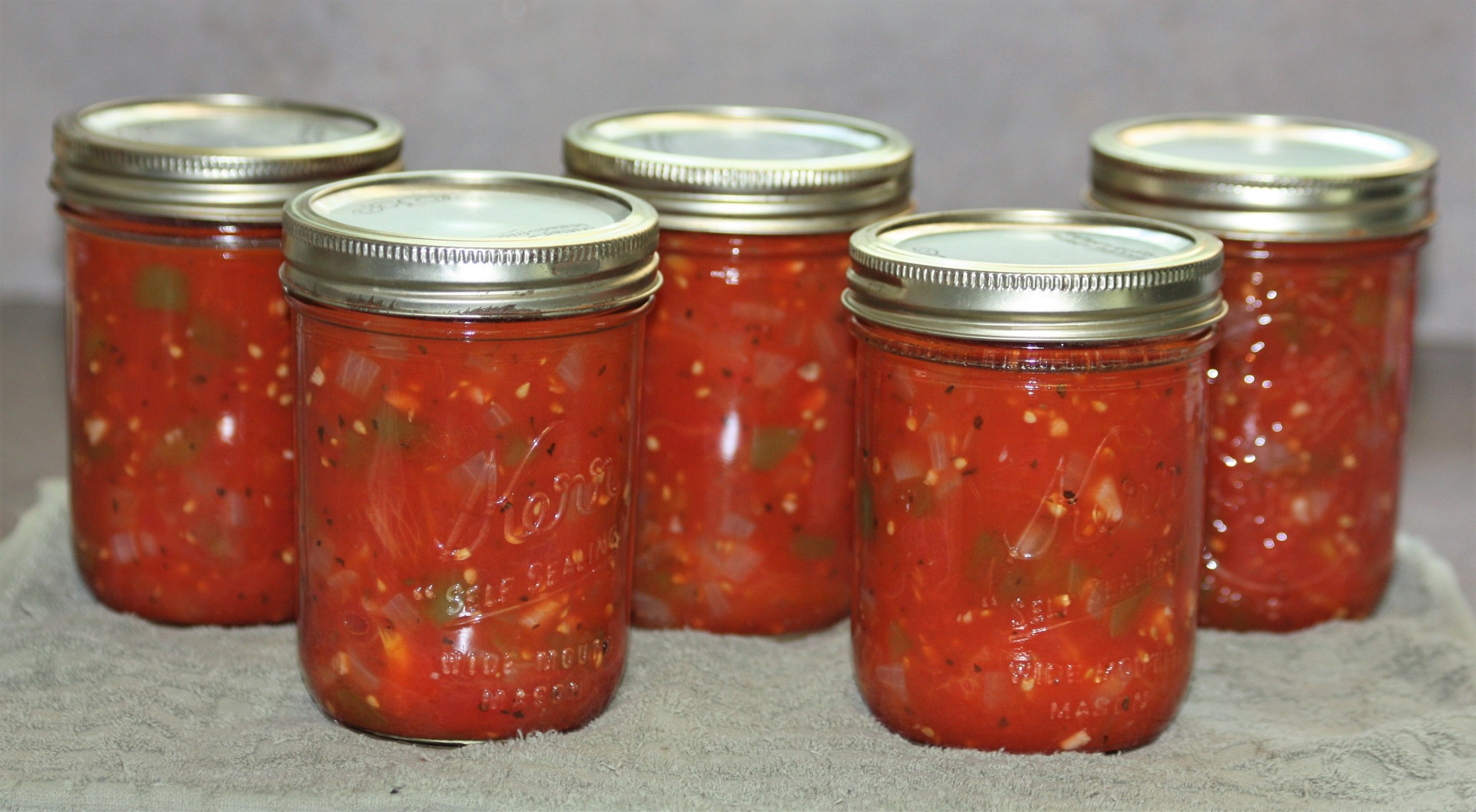Five jars of canned stewed tomatoes sitting on a gray kitchen towel with a gray background.