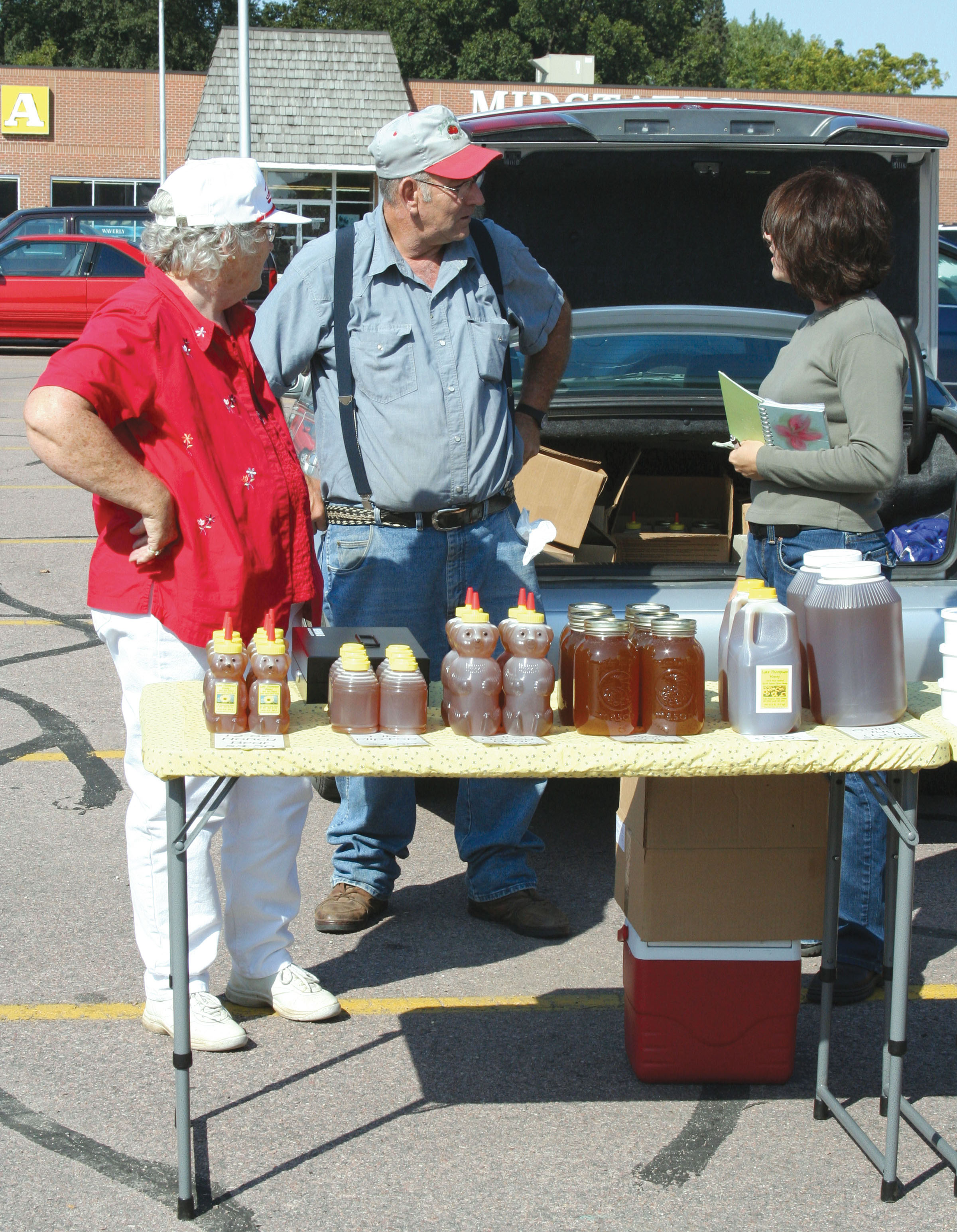 An older husband and wife talking with a female vendor near a table displaying honey products at a farmers market.