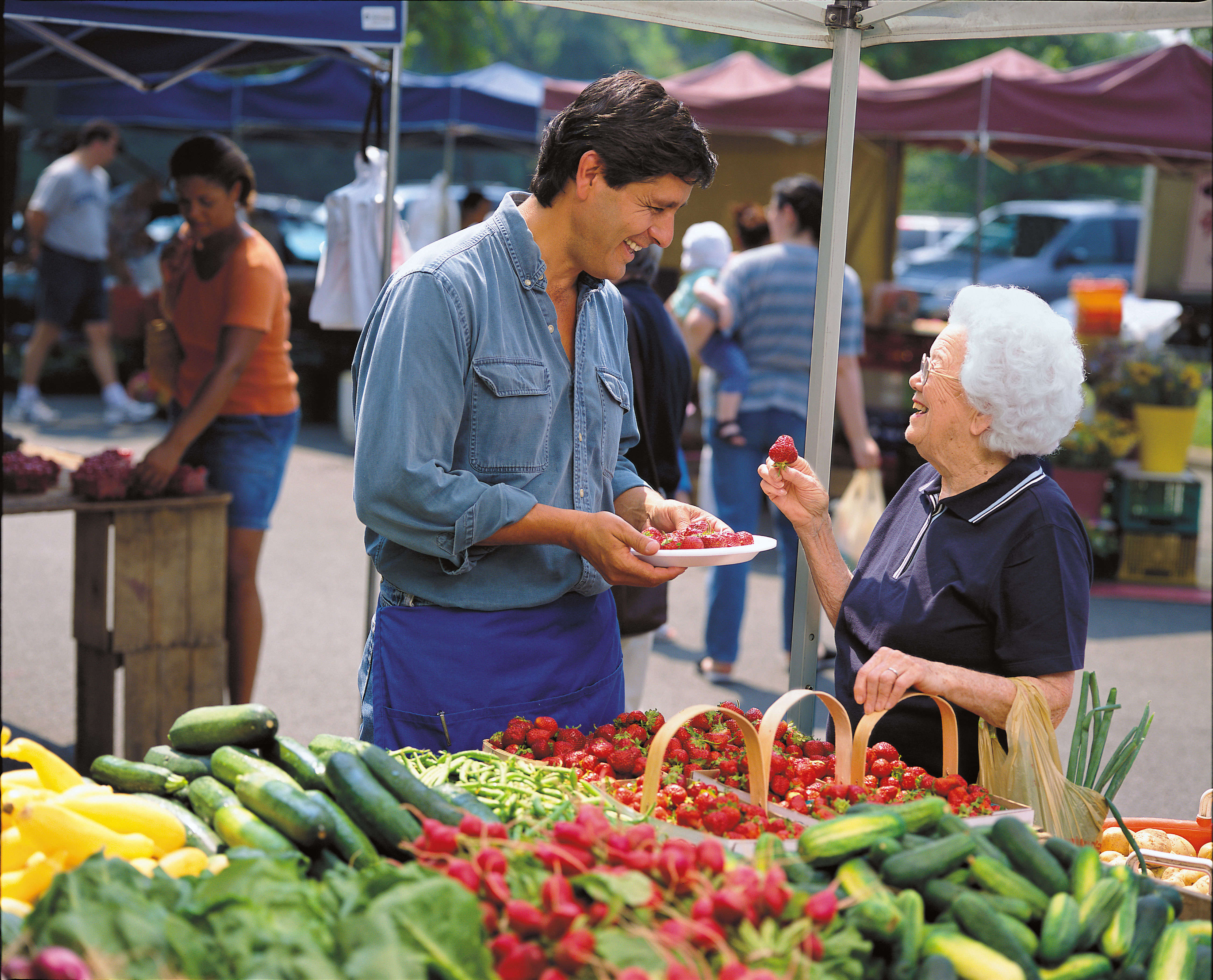 A male vendor serving a strawberry sample to an older woman at a farmers market stand.