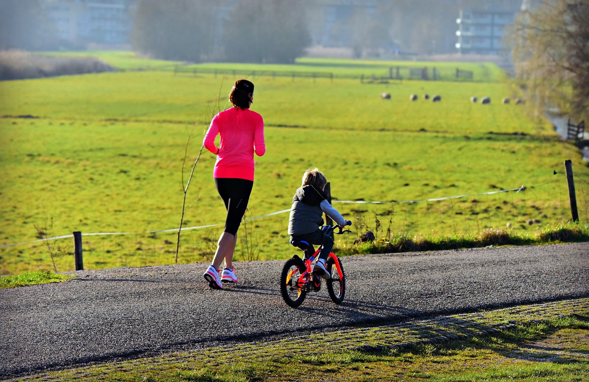 A mother jogging down a paved country trail next to a child riding an orange bicycle.