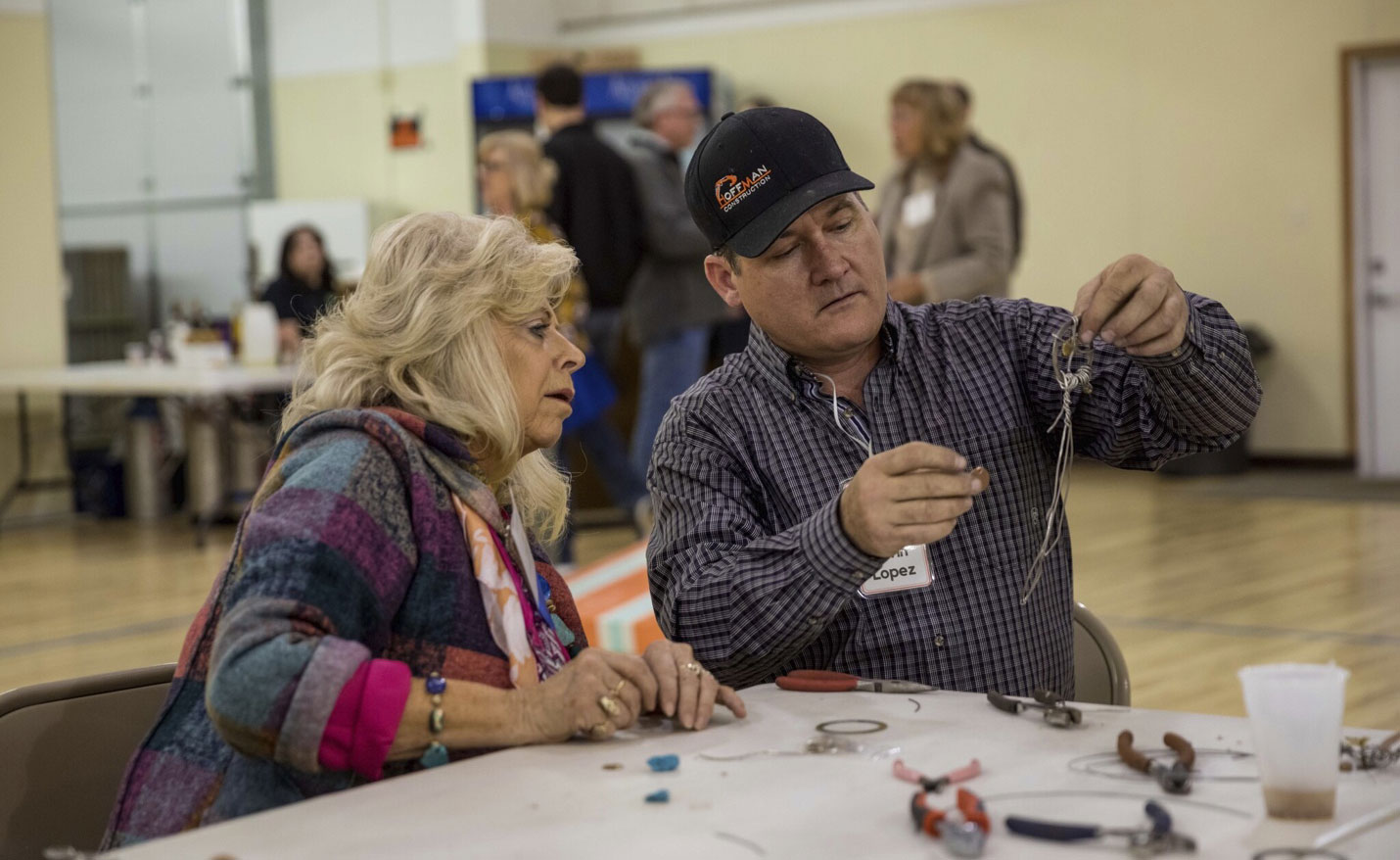 A male conference attendee in a black cap constructing a homemade dream catcher. A woman sitting next to him is closely watching.