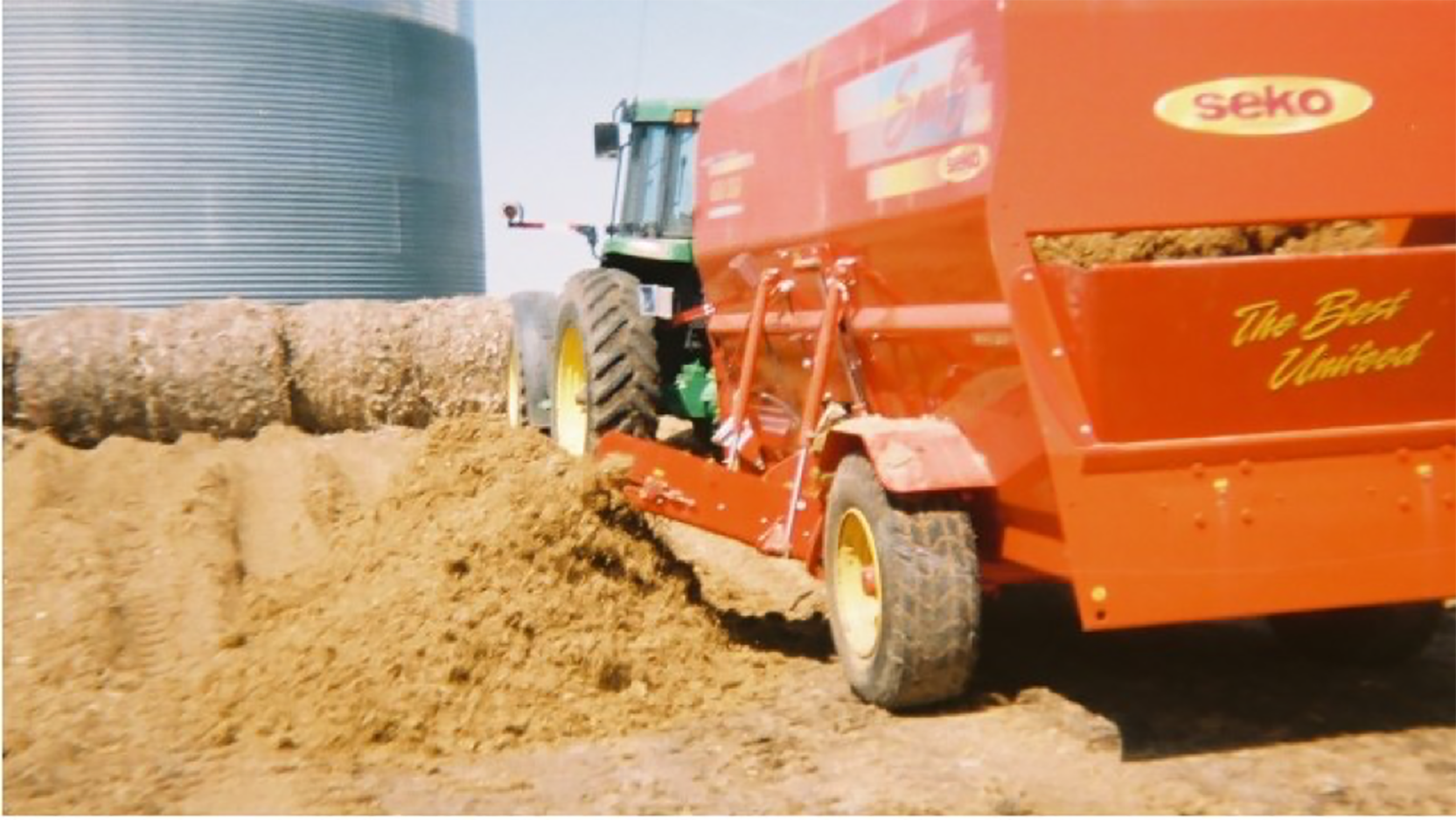 A green tractor pulling a red wagon next to a pile of wet distillers grains.