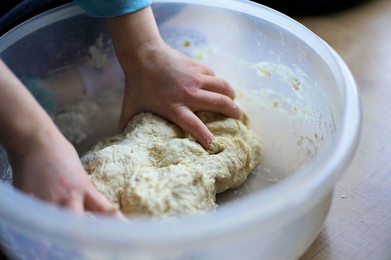 A pair of hands kneading a hydrated dough inside a plastic mixing bowl.