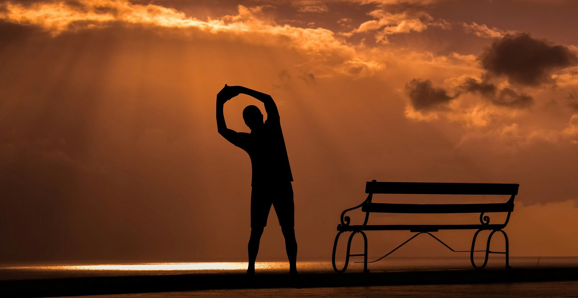 person stretching in front of the rising sun near a bench on the beach