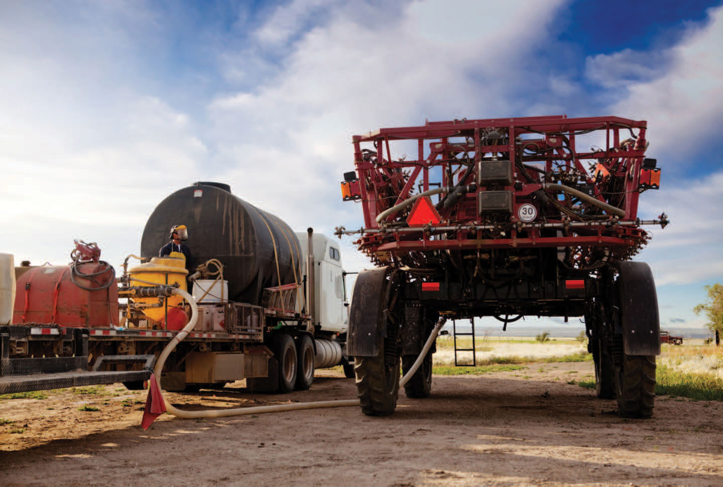 A red, tractor-mounted sprayer with its tank being flushed by a hose connected to a large truck.