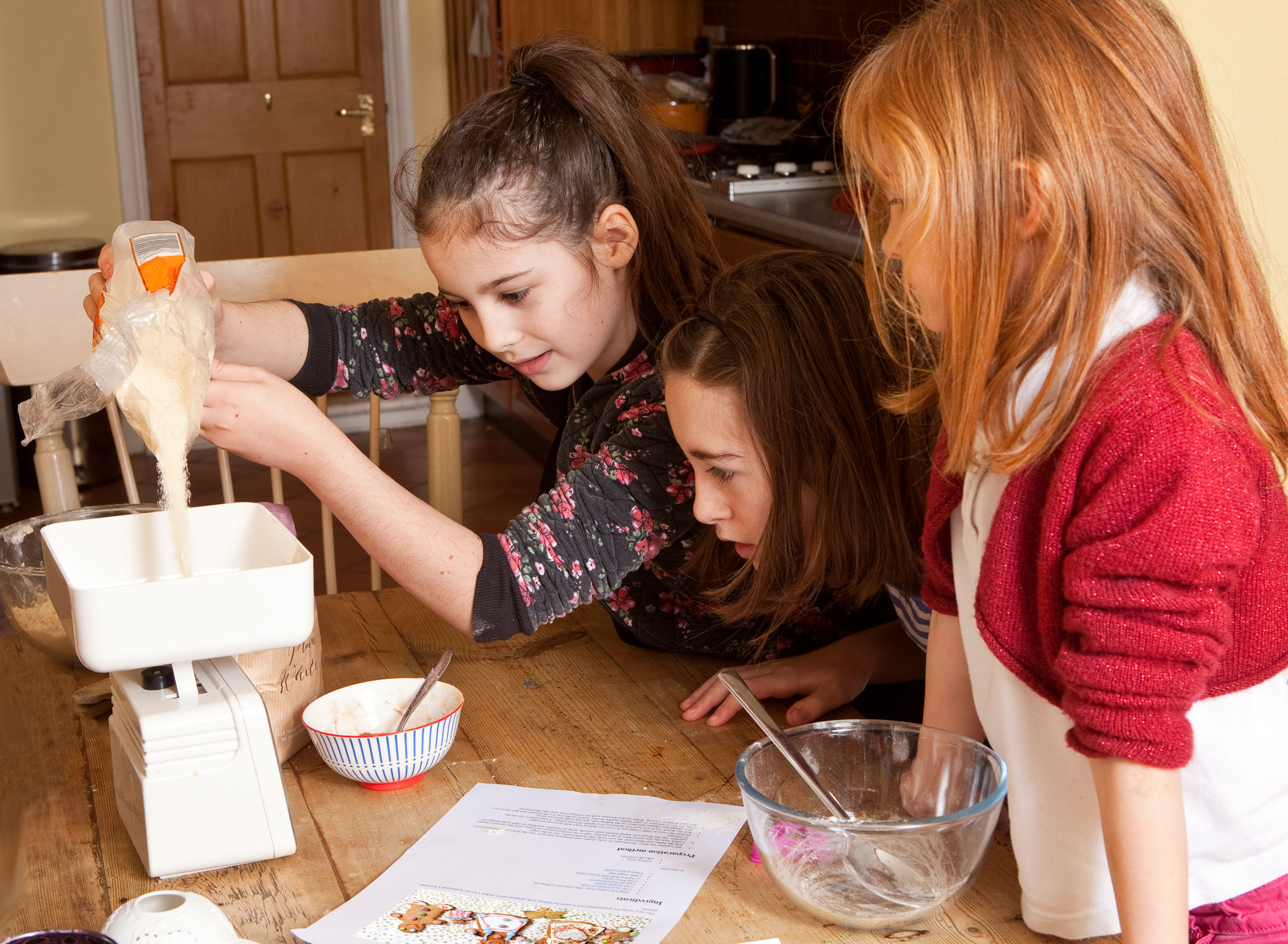 Three young girls weighing sugar on a kitchen scale.