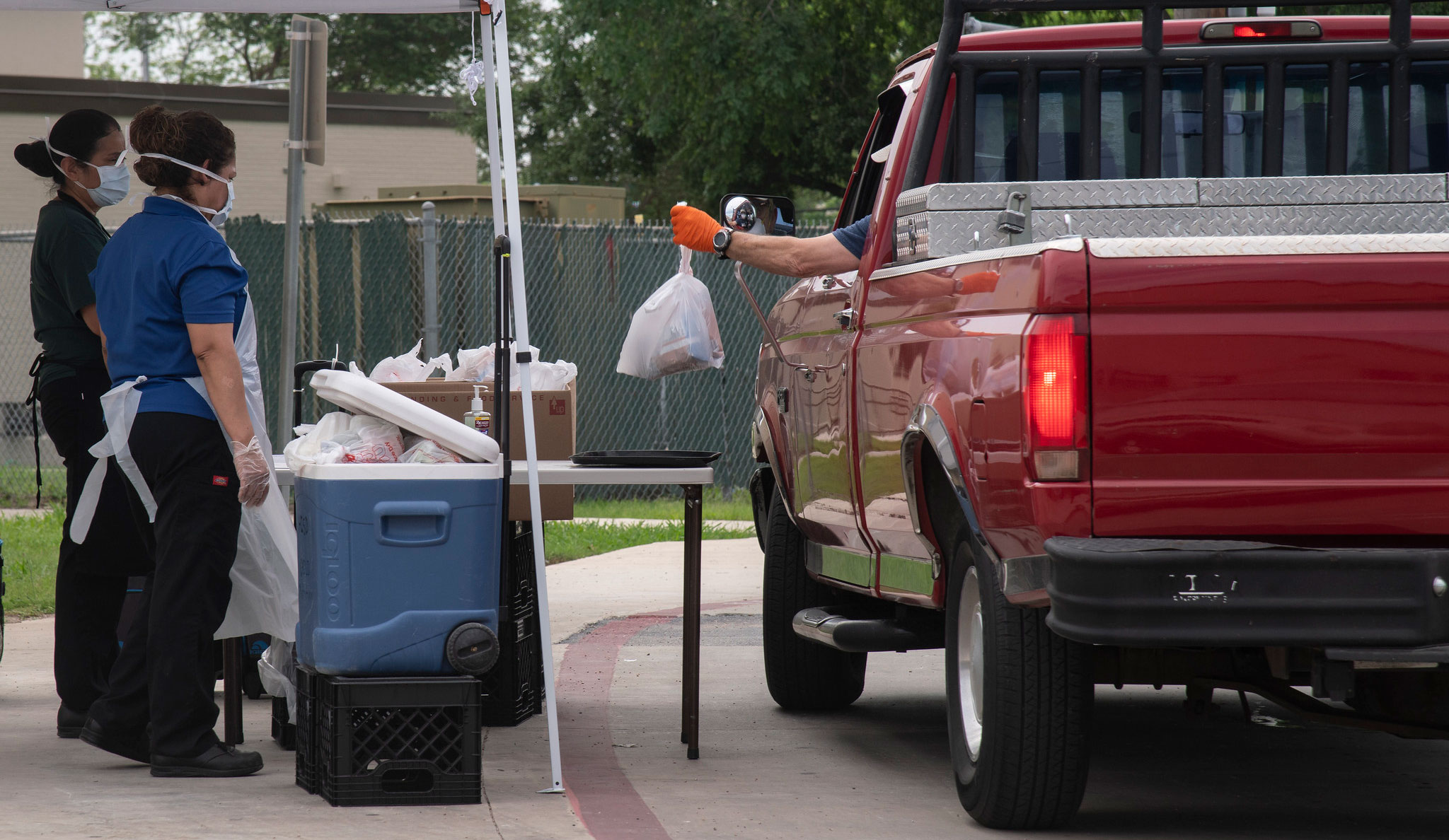 A man in a pickup truck picking up a bag of farmers market food at a pickup table being serviced by two women in personal protective gear.