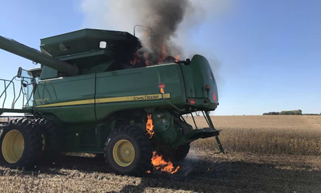 A green combine that has caught fire in the field.