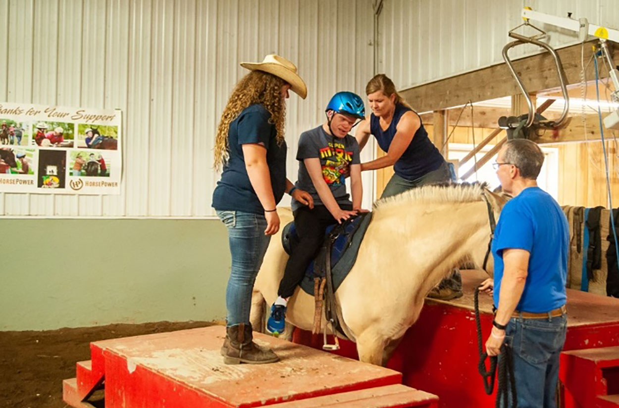 Staff of the Horse Power organization helping a young male aboard a horse for a therapeutic ride.