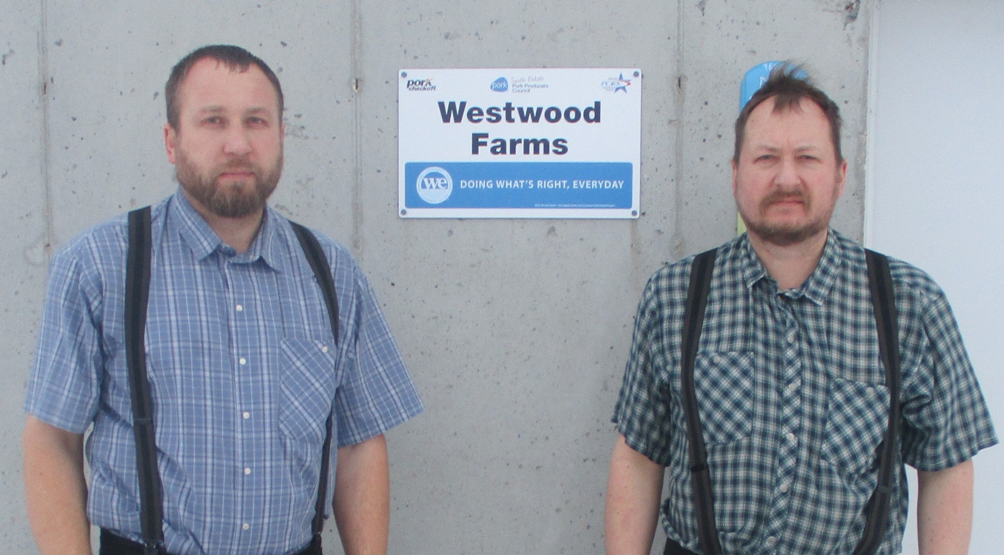 Two men standing in front of a wall with a sign hanging on the wall.