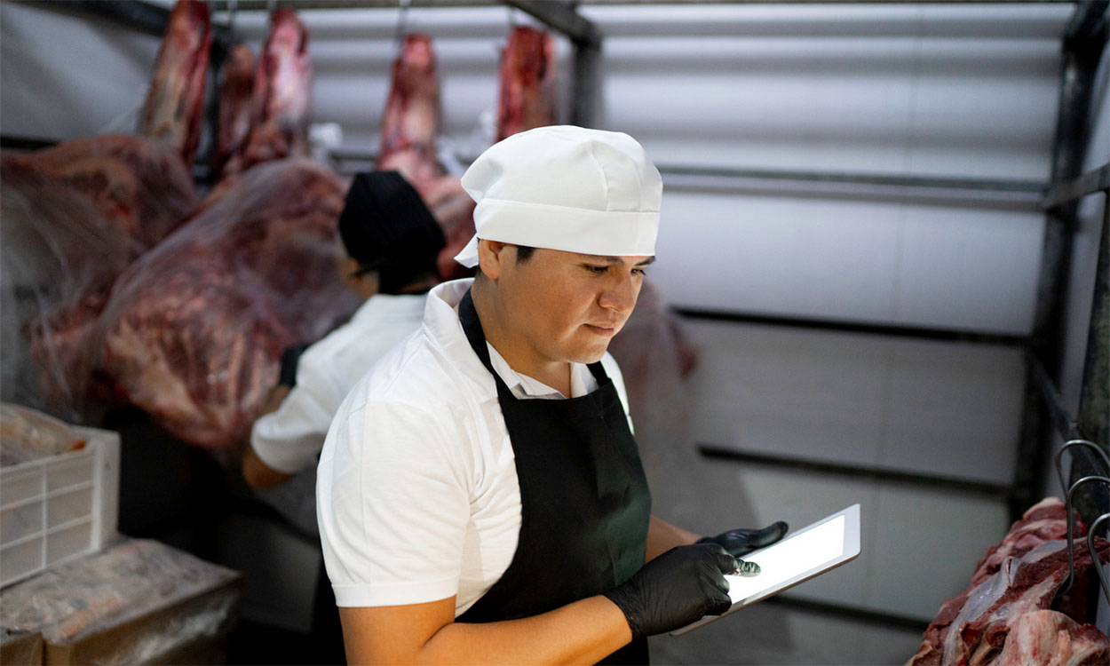 Young producer inspecting meat in a locker with a digital tablet.