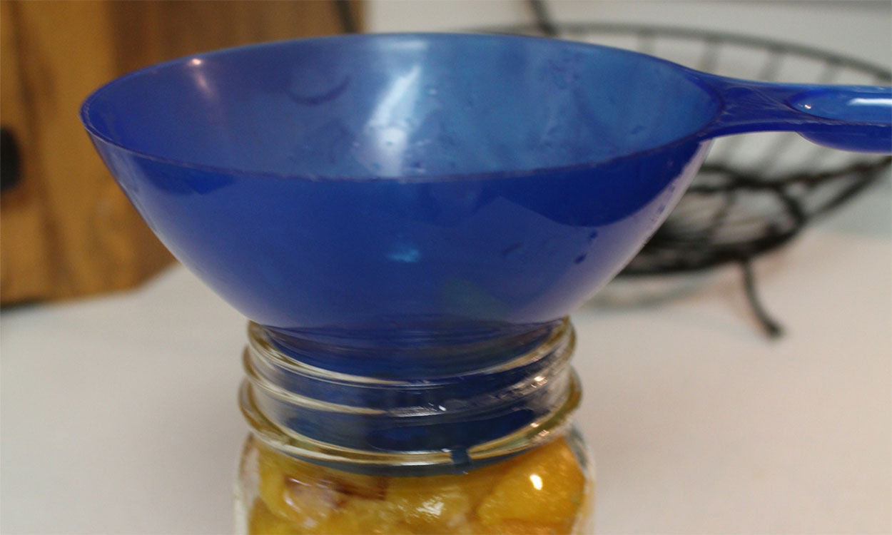 A blue funnel being used to pack syrup into a jar of peaches that are ready for canning.