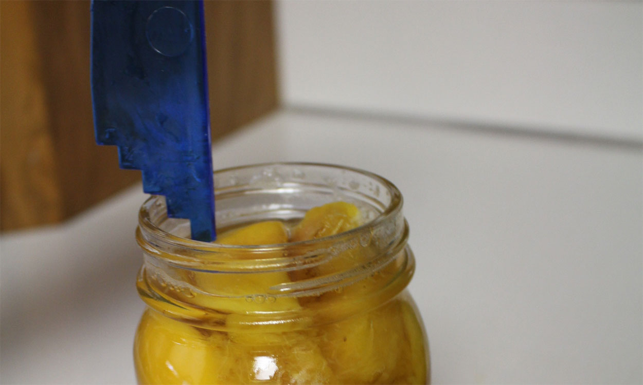 A blue measuring tool being used to measure headspace in a jar of peaches that are ready for canning.