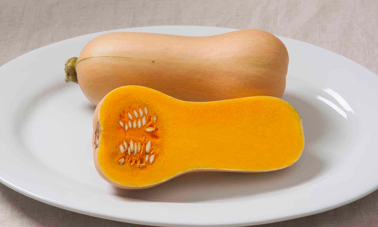 Tan, elongate squash on a white plate split open to reveal a dense, orange fruit inside.
