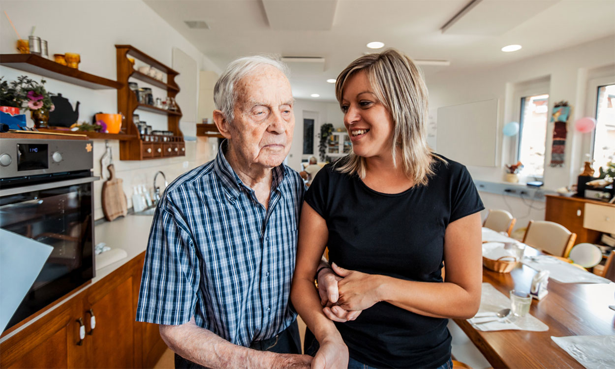 Older adult man getting assistance from a young, female caregiver.