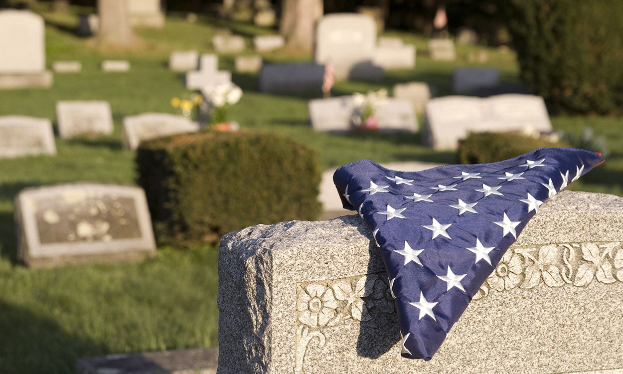 A picture of a tombstone in a cemetery with an American flag laying on it.