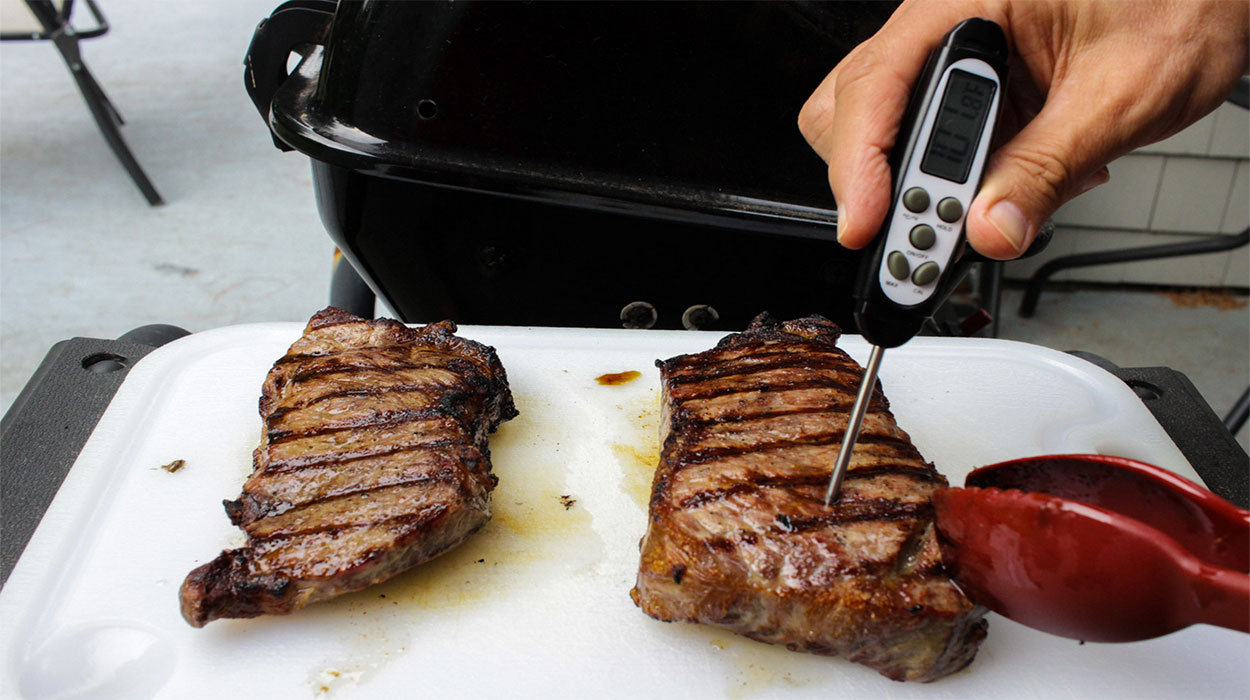 Hand holding meat thermometer to check the doneness of grilled steaks.