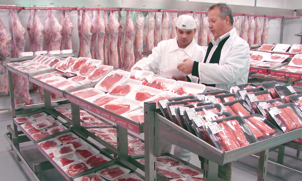 Two butchers inspecting meat cuts in a large cooler.