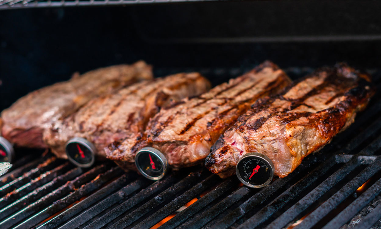 Strip steaks on grill with meat thermometers properly inserted through the side of the cuts.