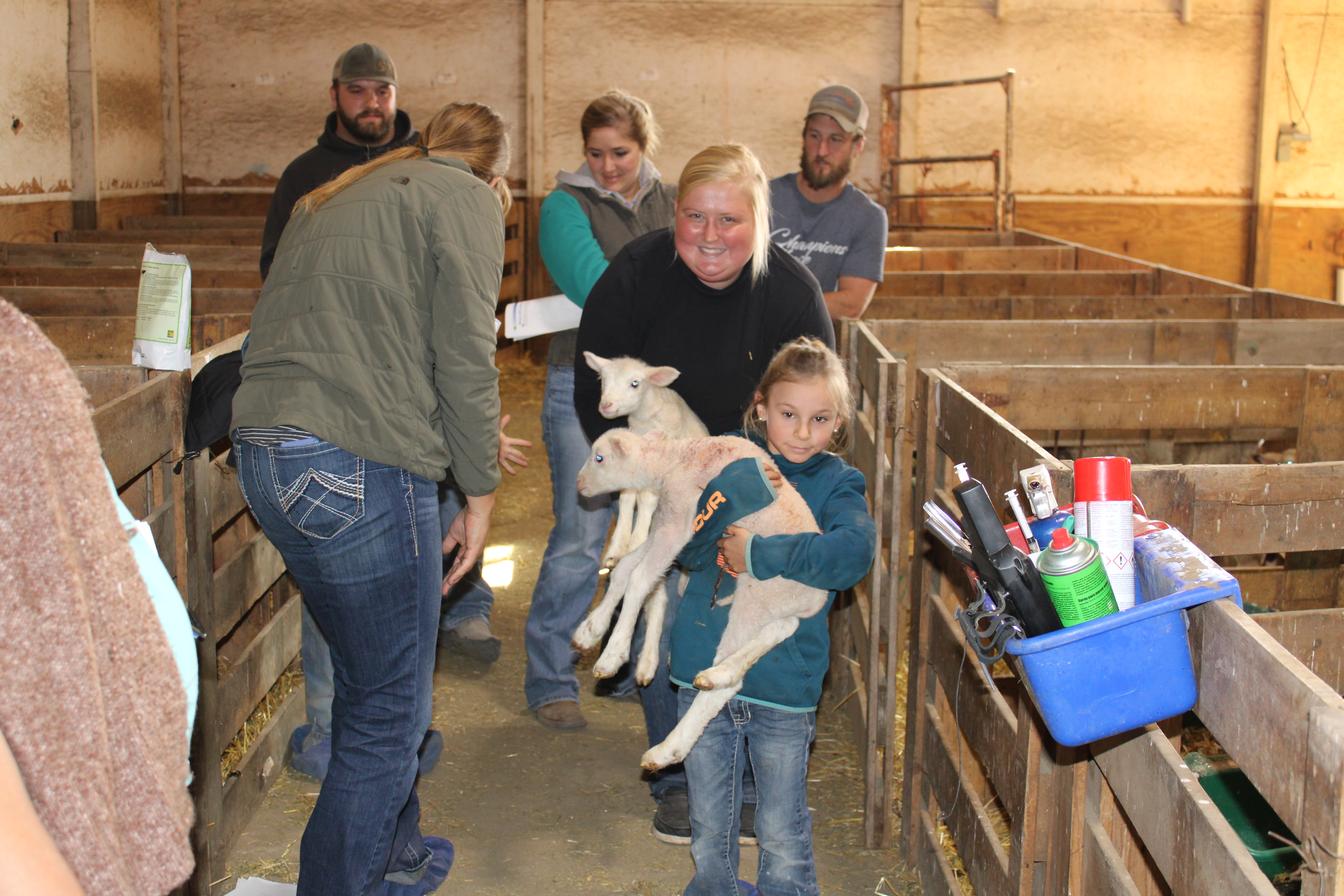 A girl holds a sheep in a barn and a blond woman stands behind her smiling