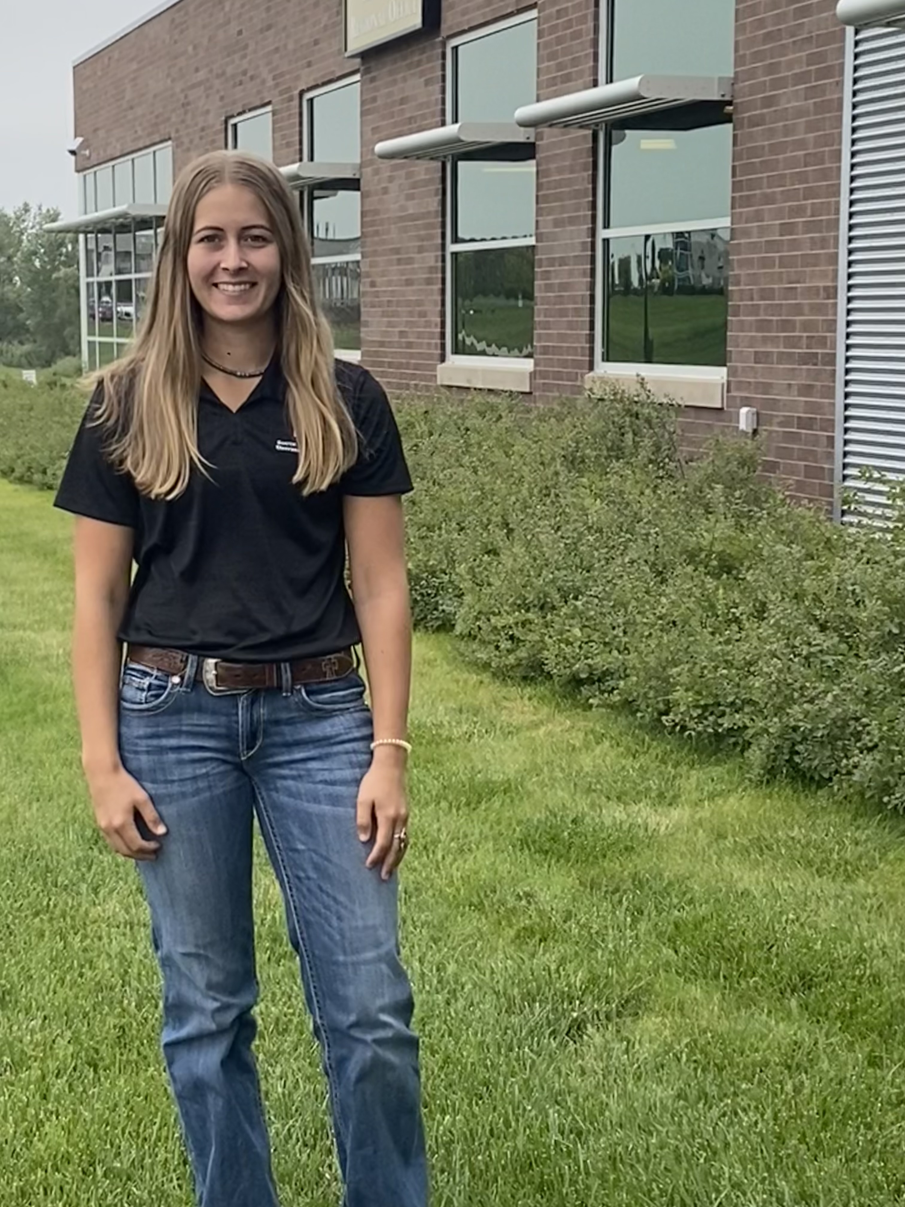 Sydney Vanderhoff stands in front of a building. Her long hair is down and she is wearing a black polo and blue jeans