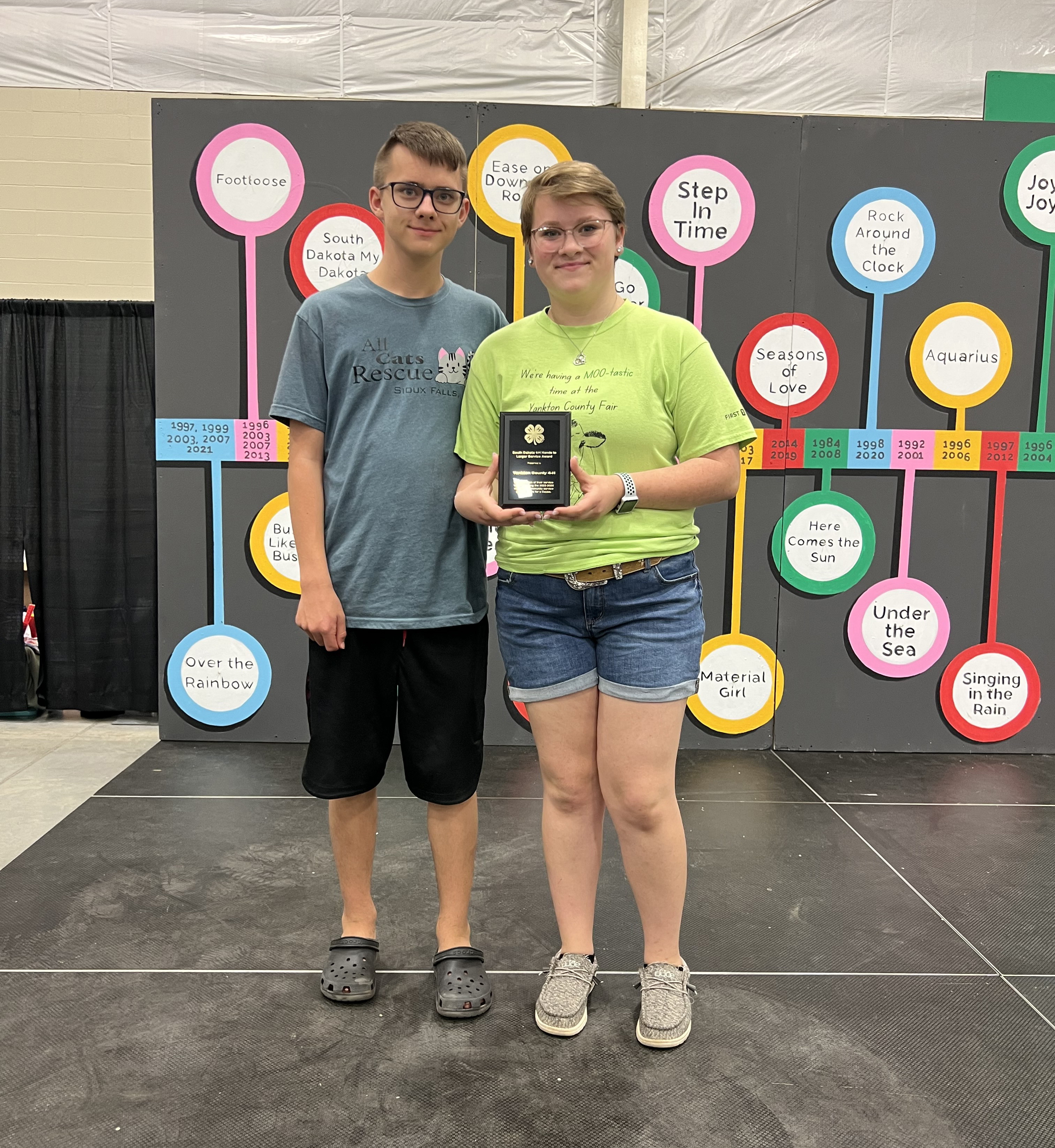 a teenage boy with short hair and glasses wearing a grey t-shirt and shorts stands next to a teenage girl with short hair and glasses wearing a bright green t-shirt and jean shorts holding an award plaque