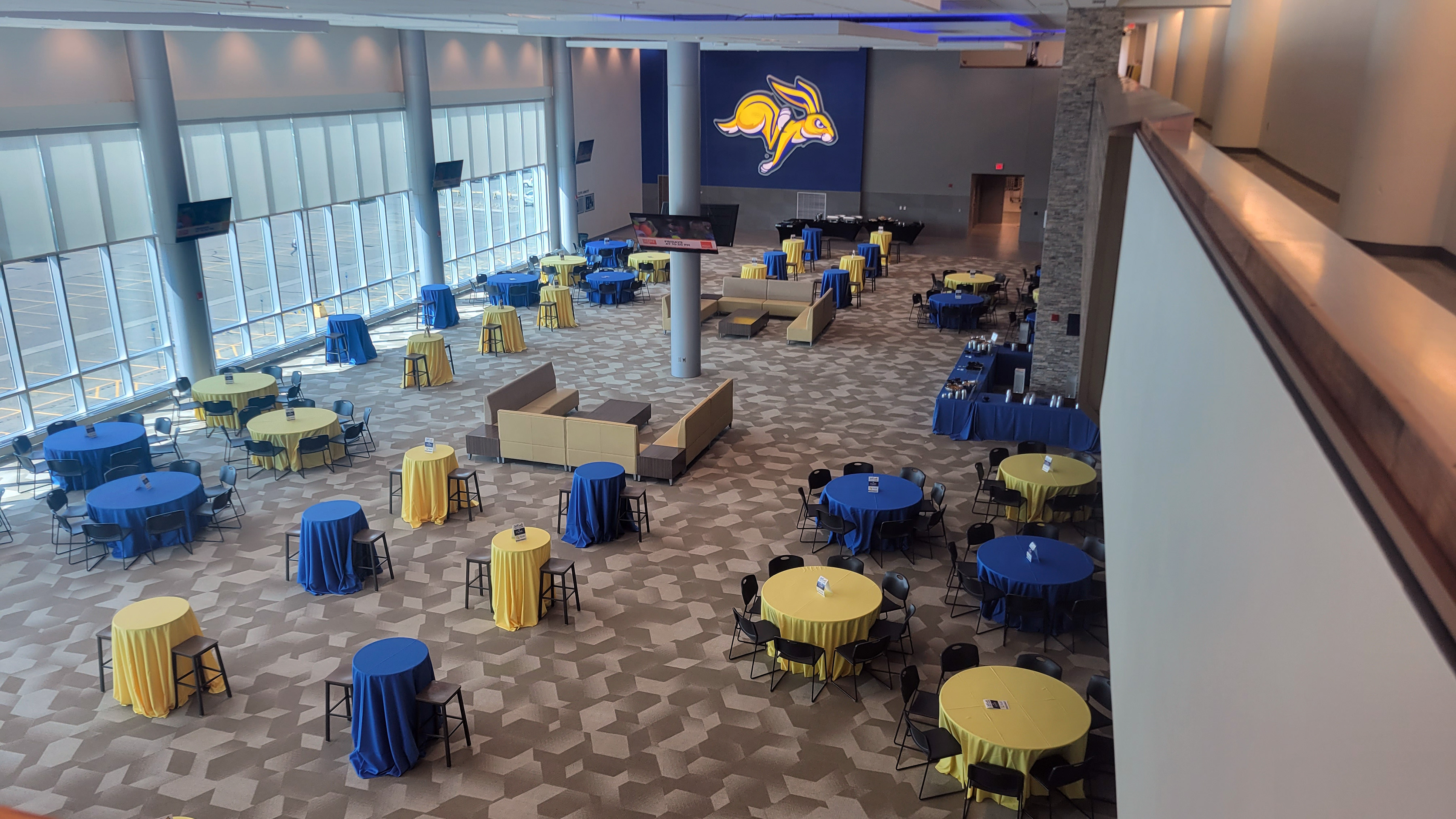 a room is pictured from above with round tables covered in blue and yellow tablecloths. a large picture of the SDSU jackrabbit logo is on the far wall