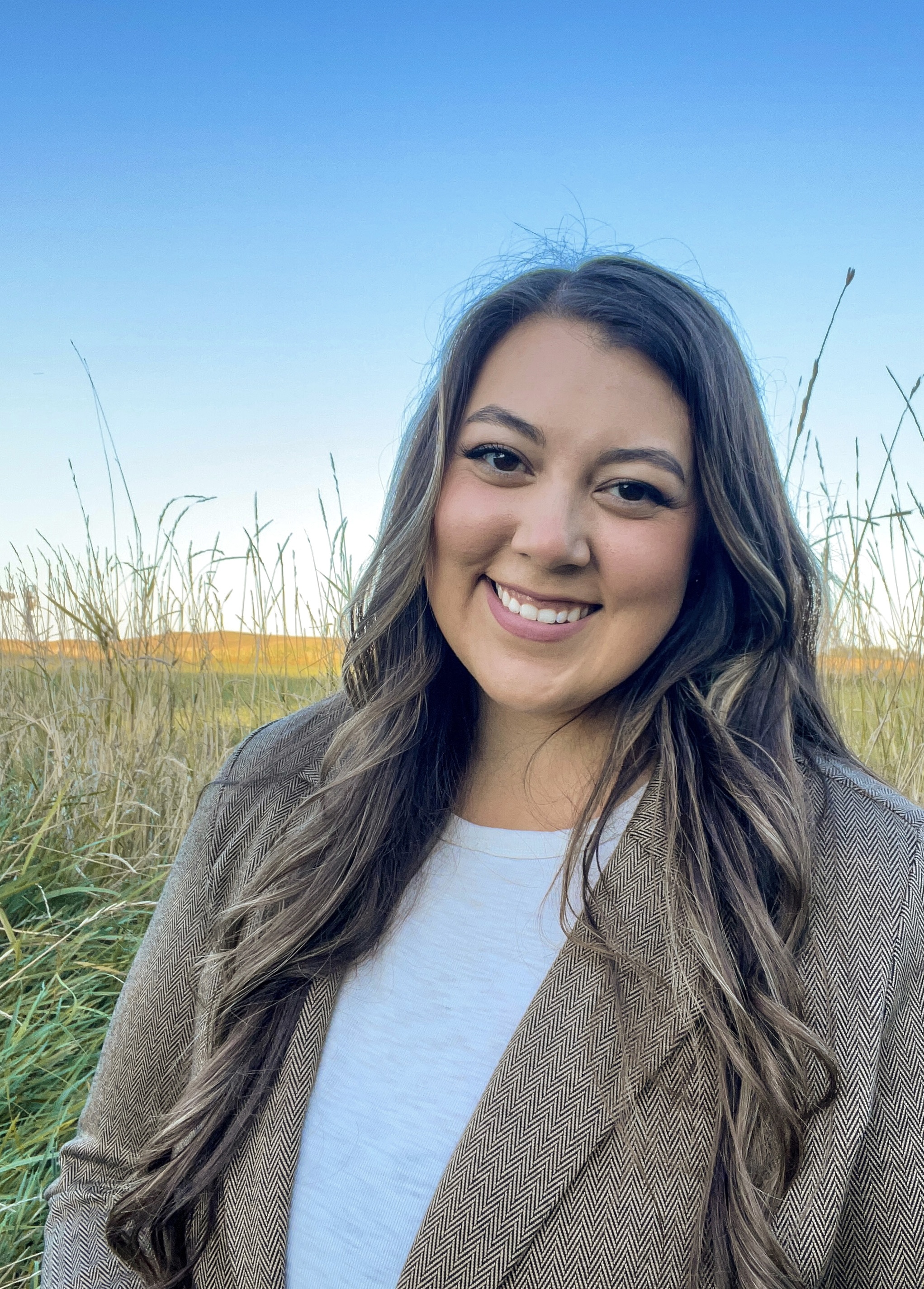 Kaylee Wheeler smiles at the camera. Her long brown hair is down, and she is standing outside in a field