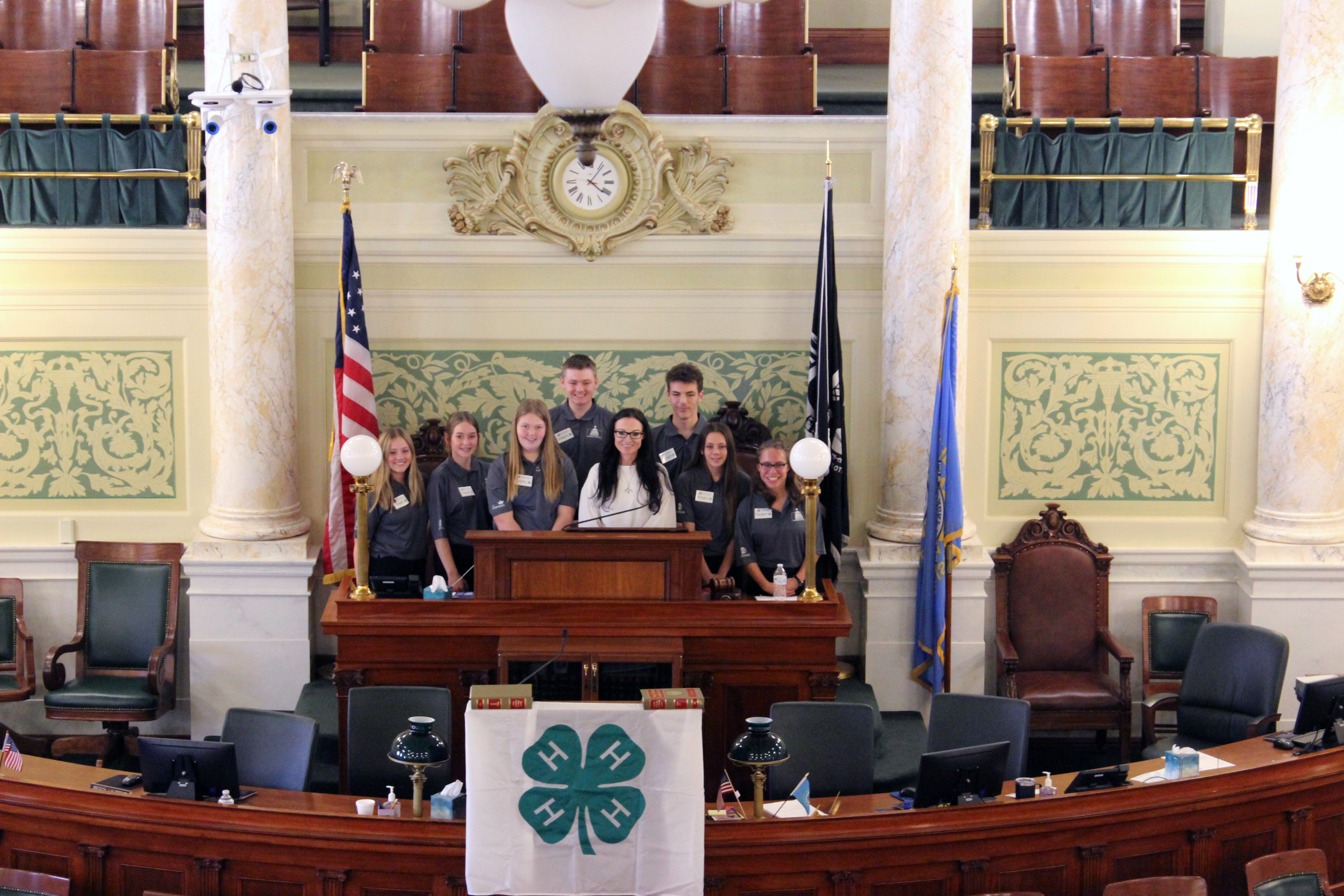 Nine youth stand behind a podium inside the South Dakota Capitol session chamber. A 4-H flag is draped in front.