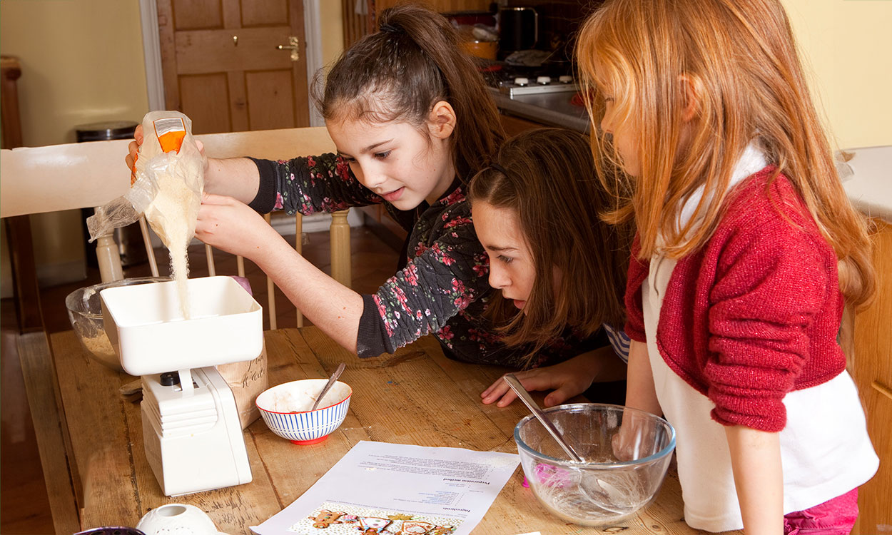 Three young girls weighing sugar on a kitchen scale.|