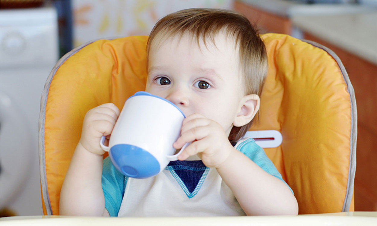Infant boy seated in a high chair drinking from a sippy cup.