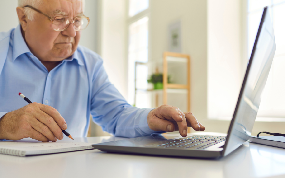 An older adult male holds a pencil over a notepad in one hand, while typing on a laptop with the other