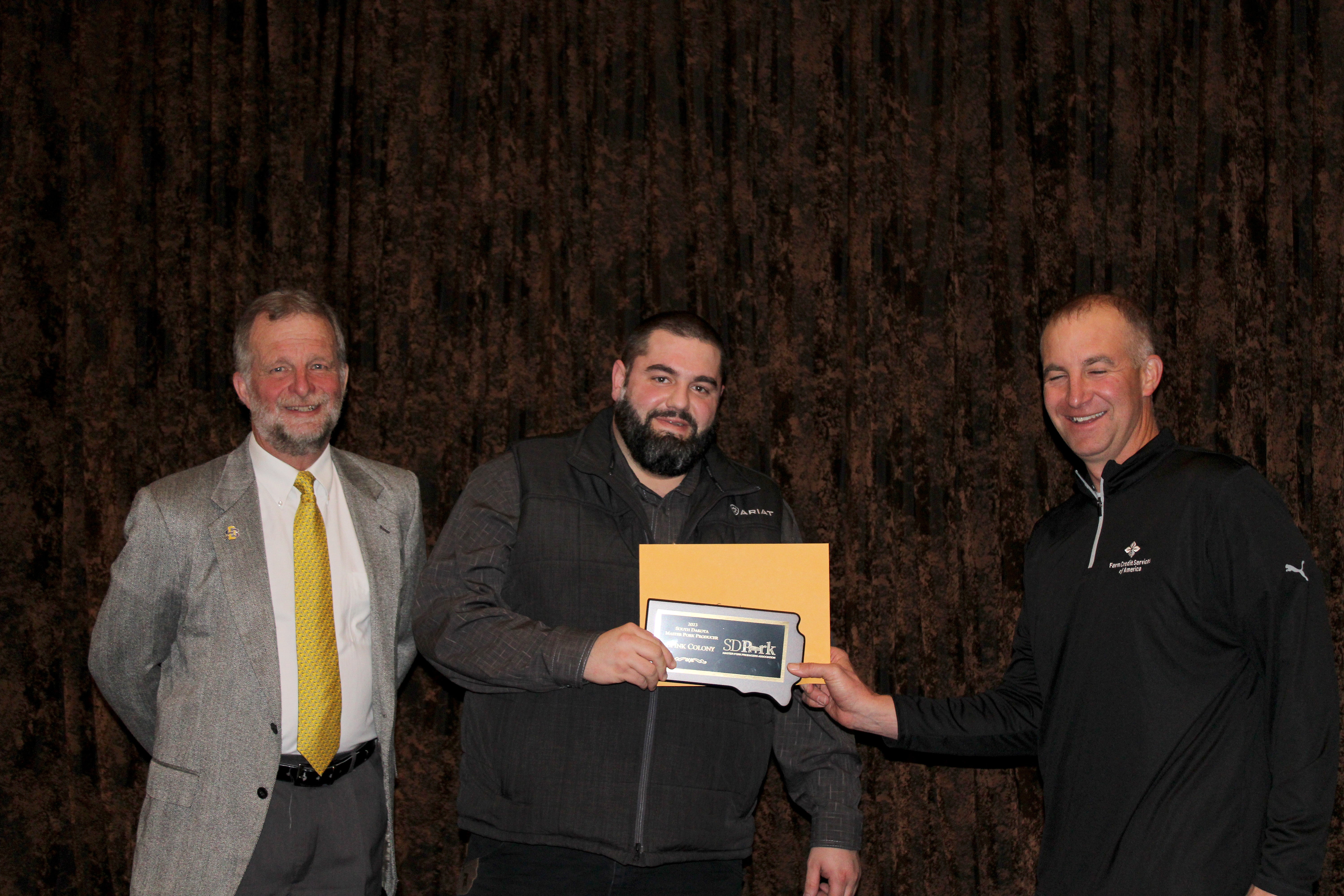 Three men smile at the camera. the man in the middle holds up a plaque in the shape of South Dakota