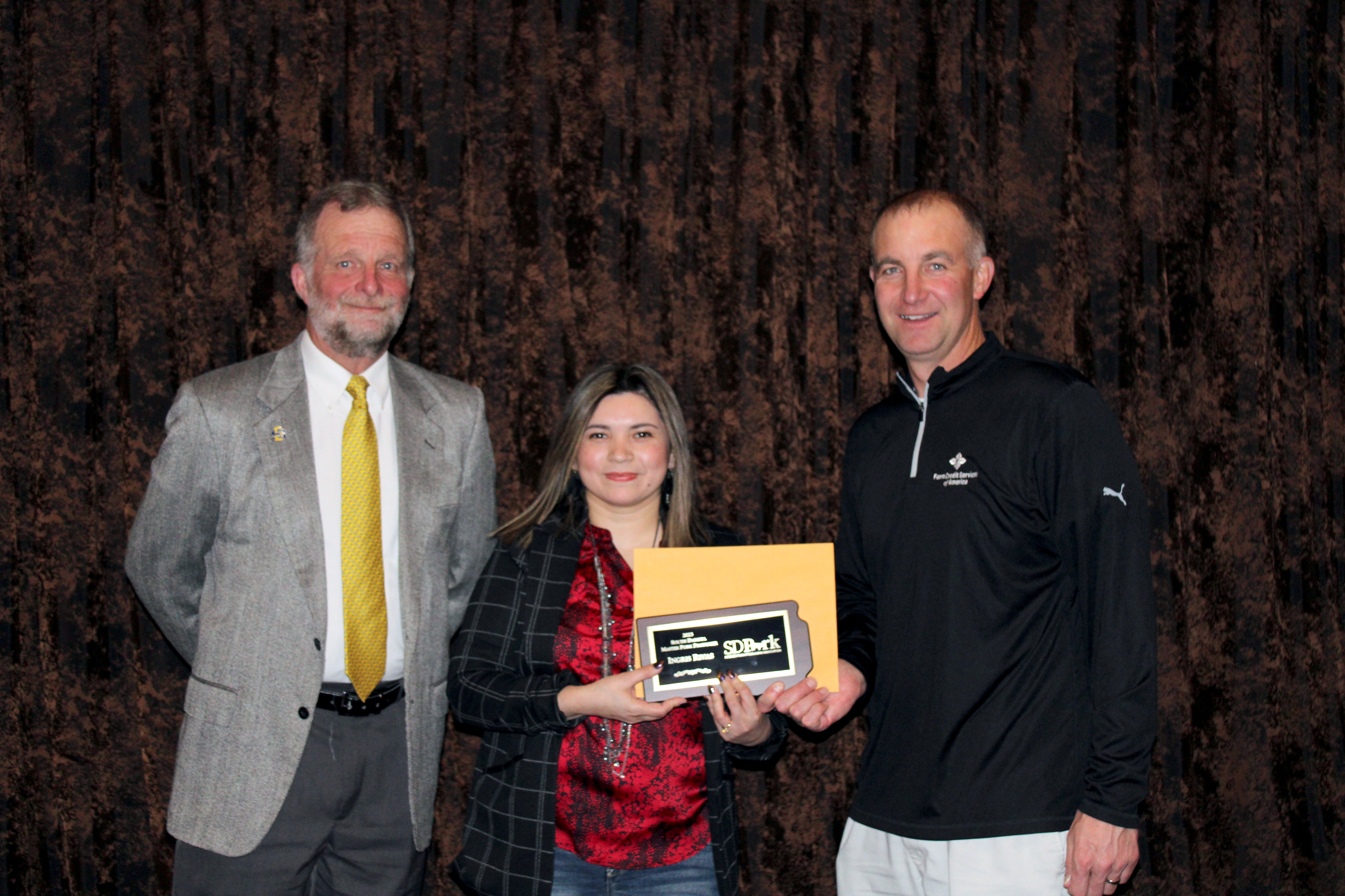 Ingris Rivas stands in between two men. She is holding a plaque in the shape of South Dakota