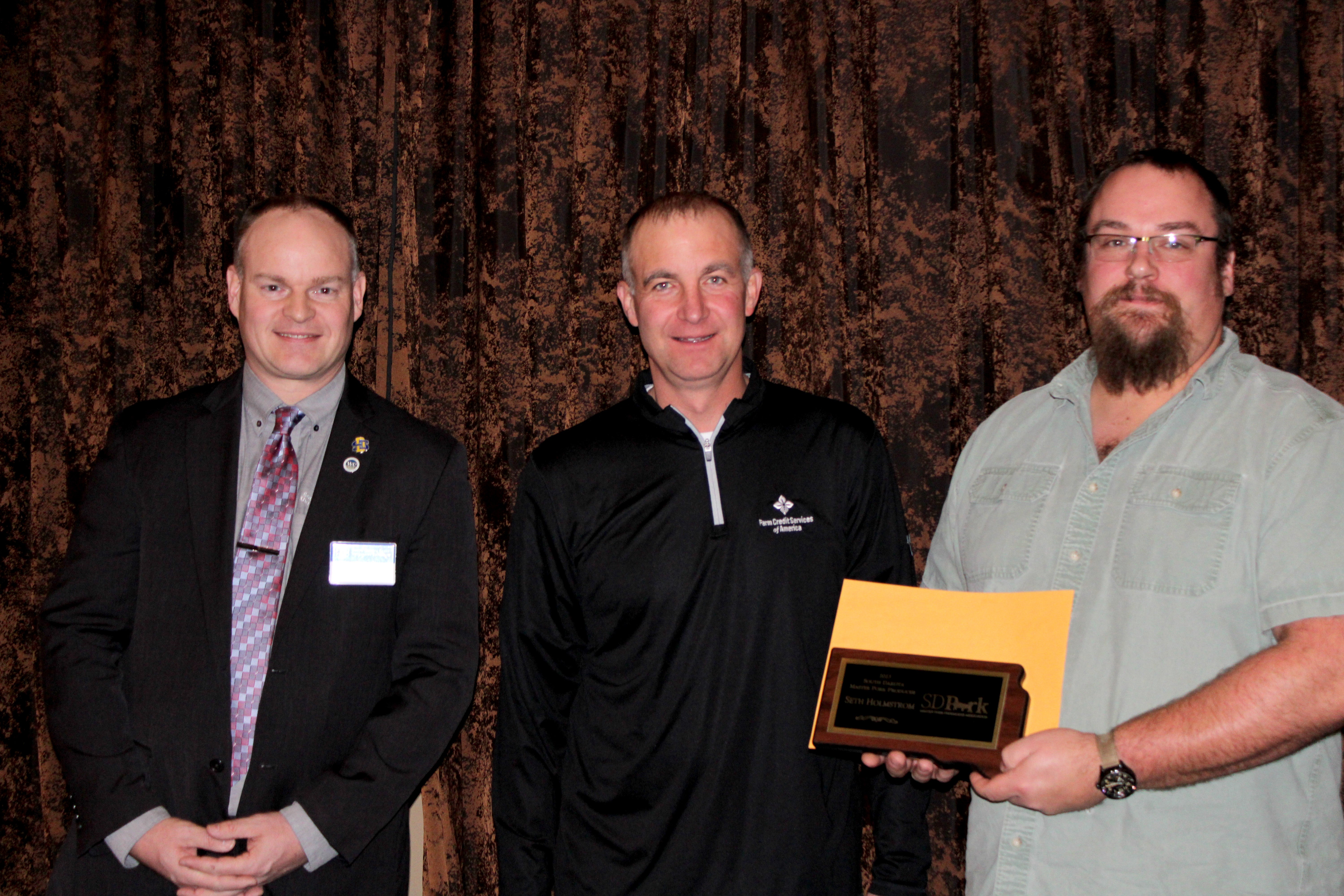 Seth Holmstrom stands next to two men facing the camera as Holmstrom holds up his plaque