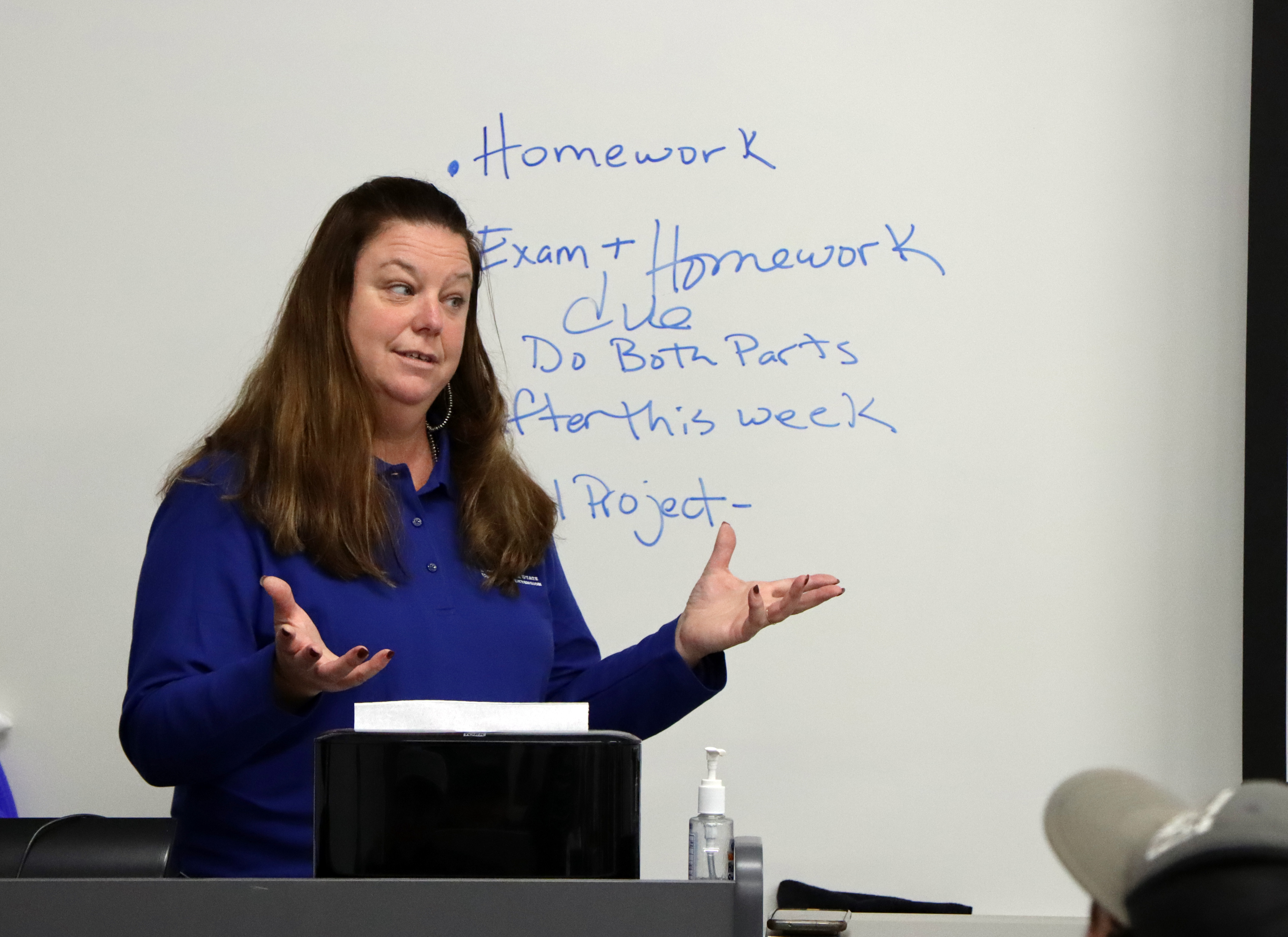 Heather Gessner gestures to her classroom. She's wearing a blue shirt and standing in front of a white board with the bullet points homework, exam plus homework due, do both parts, after this week