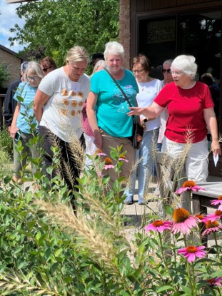 A woman in a red shirt leads a tour group through a garden. There are tall grasses and pink flowers in the foreground