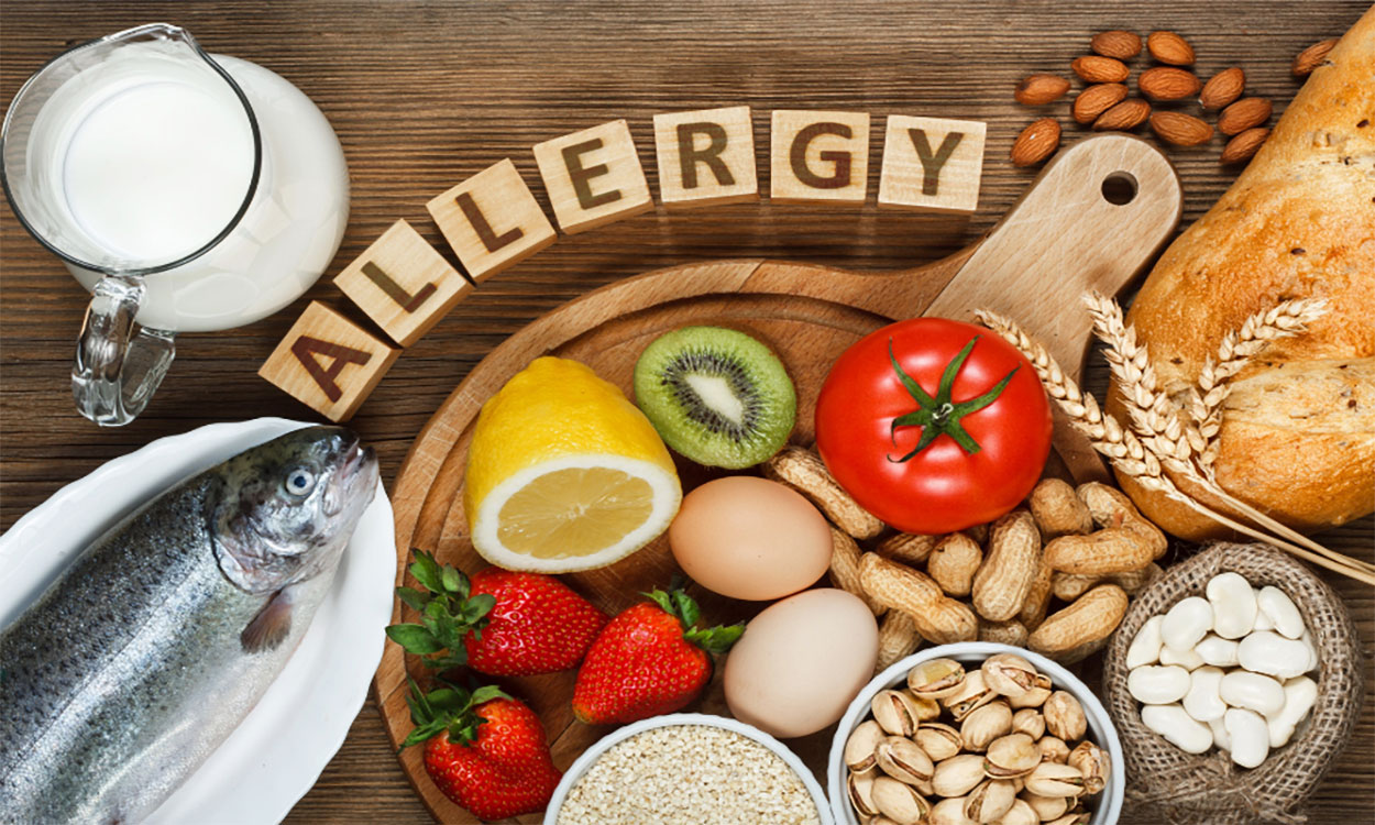 A variety of common food allergens arranged on a table surrounded by the word “Allergy” spelled out on wooden tiles.