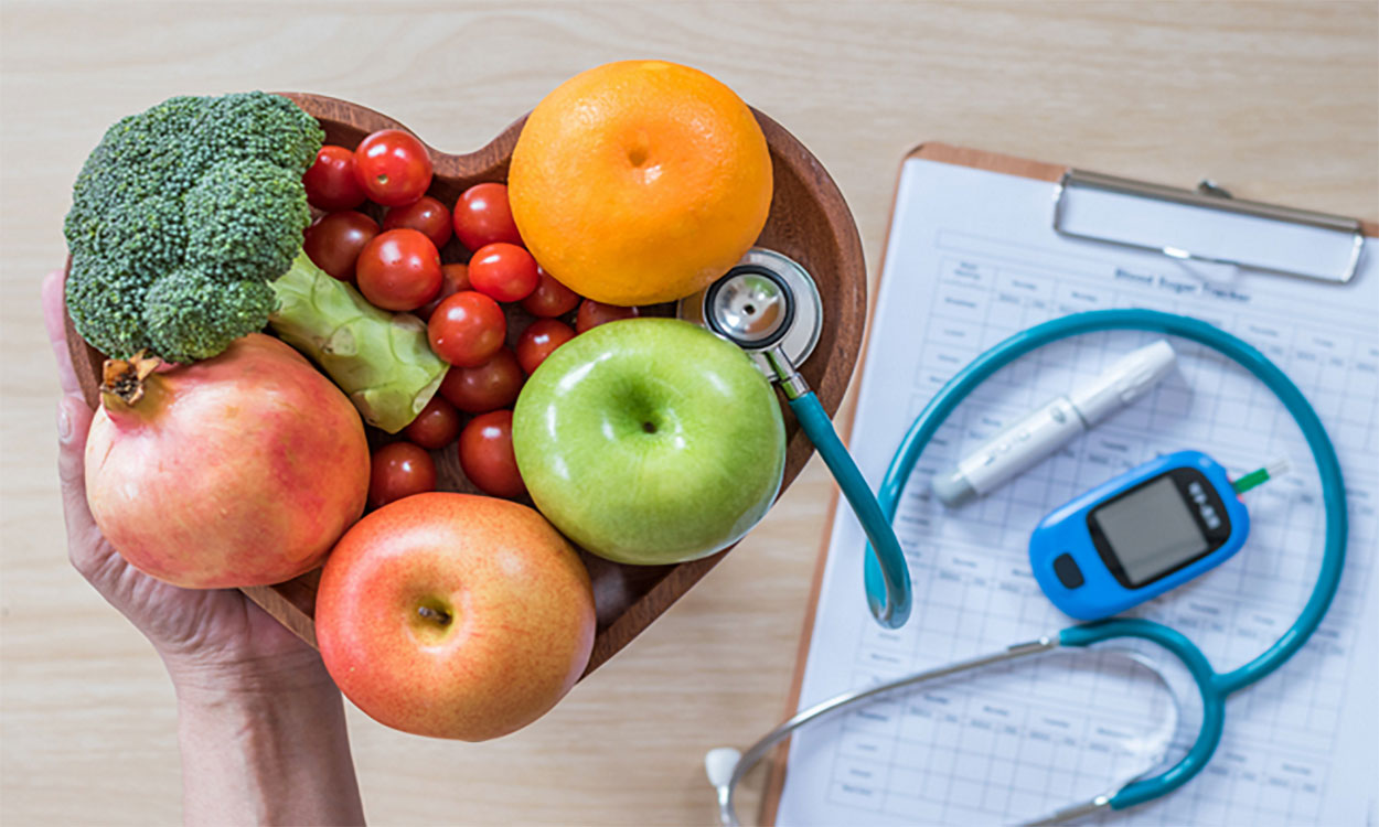 Hand holding a heart-shaped bowl of fruits and vegetables near a clipboard, stethoscope, and glucose meter.