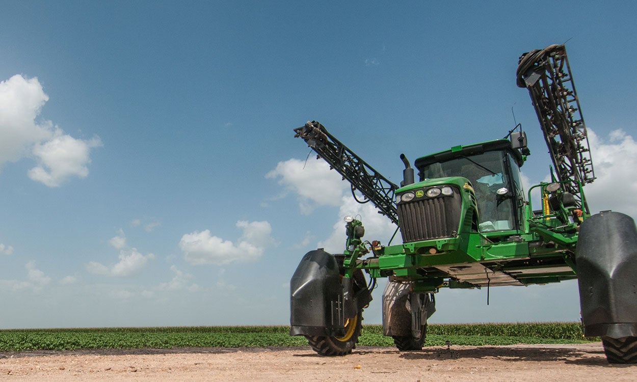 A green boom sprayer parked outside a crop field.
