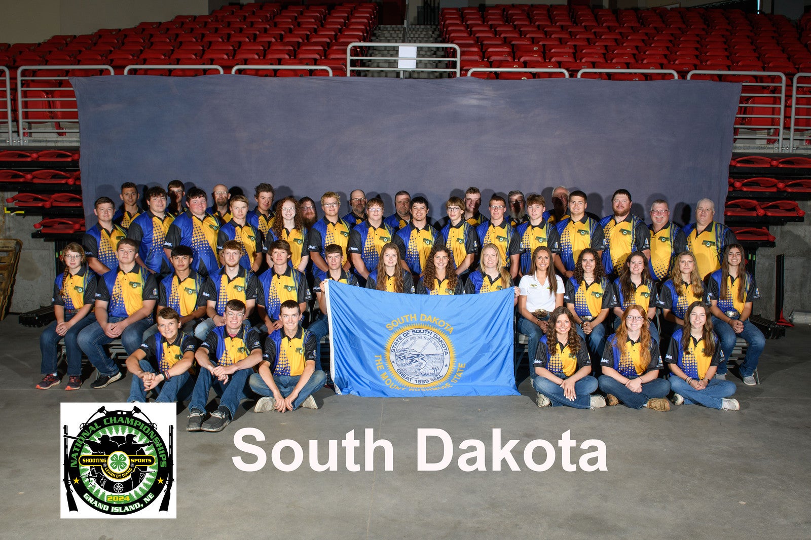 A large group of youth in matching blue and yellow shirts pose. They are holding a South Dakota flag in front of them