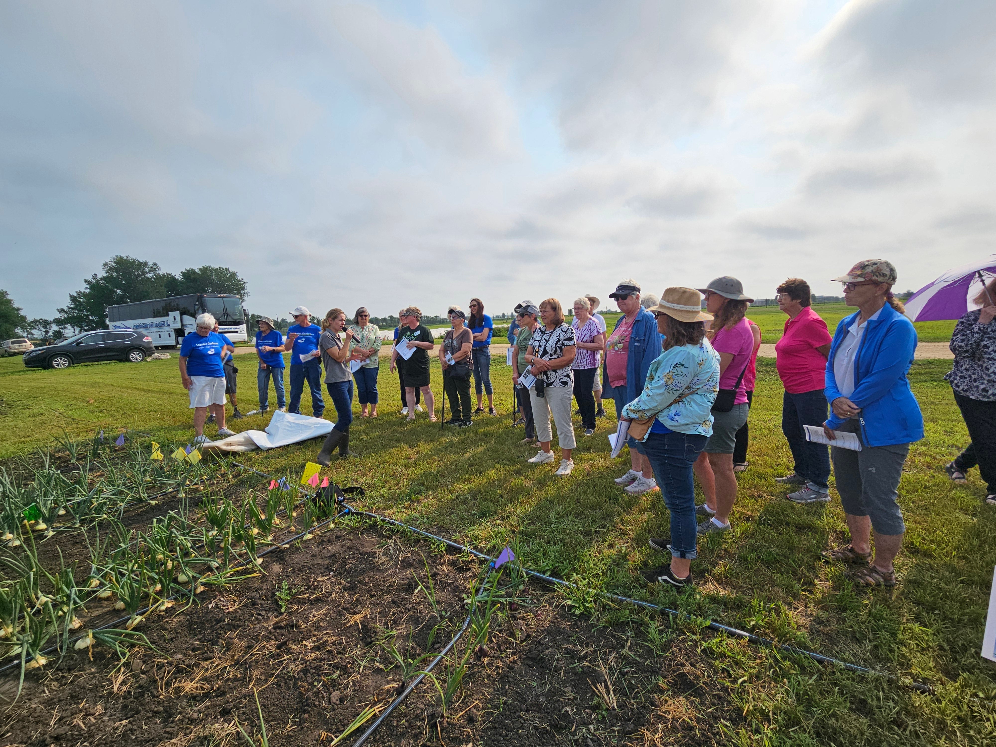 A woman with a microphone speaks to a group of people standing outside rows of planted vegetables