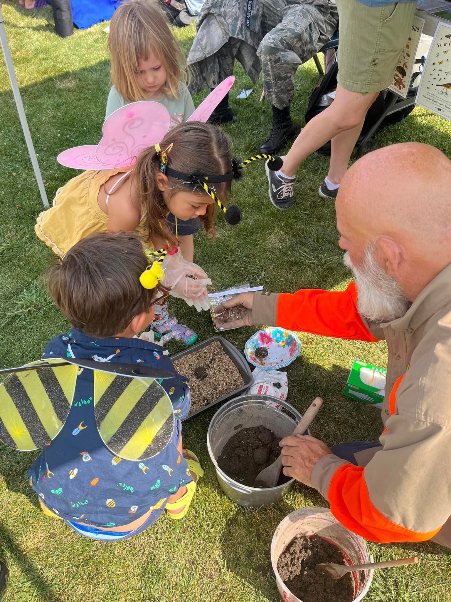 A little girl wearing butterfly wings and a little boy wearing honeybee wings scoop dirt and insects out of pails